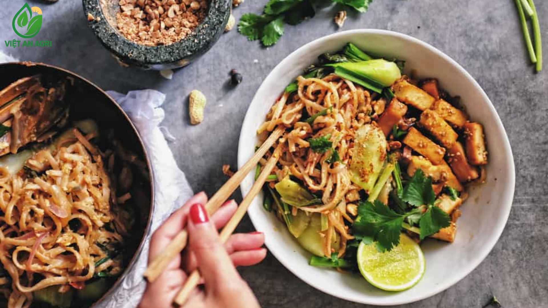 Một-người-đang-dùng-Đũa-gắp-Món-Mì-xào-kiểu-Thái-hoặc-Phở-xào-chay-với-Đậu-hũ-chiên-và-Rau-cải-trong-một-bát-sứ-trắng-có-trang-trí-Chanh-và-Rau-mùi-Cối-và-chày-đá-đen-có-Đậu-phộng-rang-giã-nhỏ-bên-cạnh-một-chiếc-nồi-khác-chứa-Mì-trên-nền-xám-đen