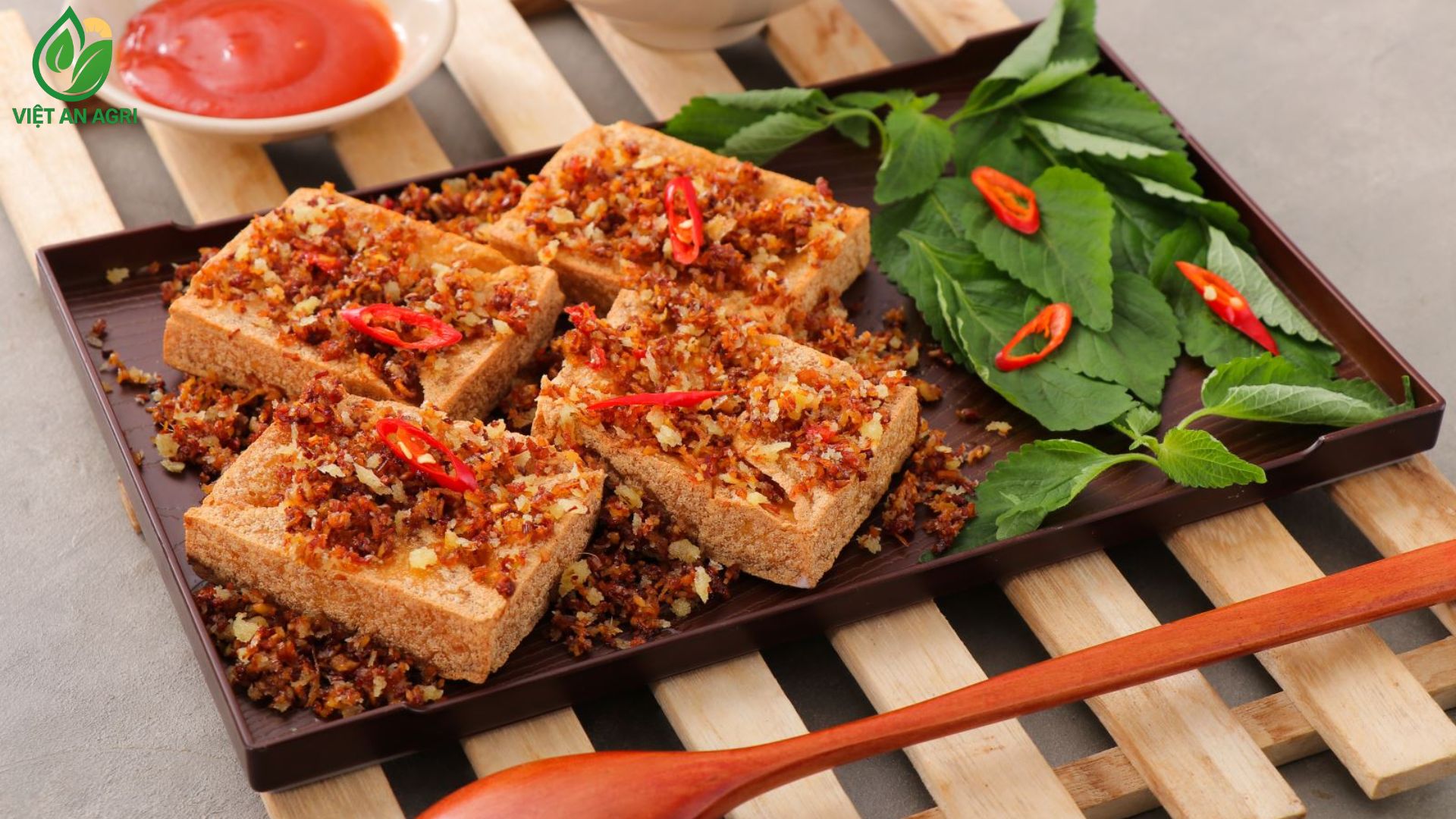 Bốn-miếng-Đậu-hũ-chiên-hình-vuông-với-Tỏi-và-Hành-khô-phi-giòn-rắc-trên-khay-nhựa-nâu-trên-các-thanh-gỗ-nhạt-Bát-sốt-chấm-cà-chua-và-Cơm-ở-phía-trên-muỗng-gỗ-dài-và-Lá-tía-tô-hoặc-Rau-húng-với-ớt-cắt-lát